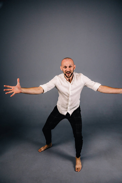 studio portrait of a bald guy with a beautiful beard barefoot in a white shirt and plaid pants dancing on a gray background - Foto, imagen