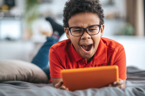 A boy in orange tshirt watching something online and yawning - Fotó, kép