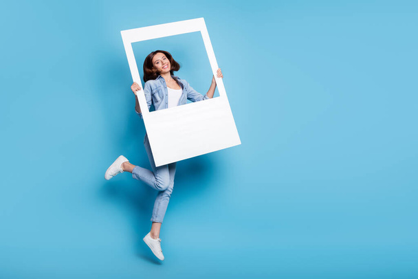 Photo of shiny cute woman wear jeans shirt holding white frame jumping empty space isolated blue color background - Fotografie, Obrázek