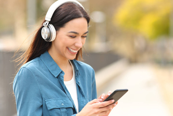 Happy woman wearing headphones listens to music or is watching media on smart phone in a park - Fotó, kép