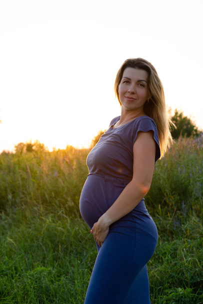 Frohe gesunde Schwangerschaft und Mutterschaft. Porträt einer schwangeren jungen kaukasischen Frau in einem langen blauen Kleid, die im Park vor der Tür posiert. Schöne weibliche werdende Dame. - Foto, Bild