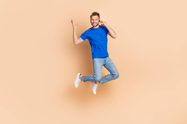 Full length photo of young man jump up tell listen talk mobile communication energetic isolated over beige color background - Foto, afbeelding