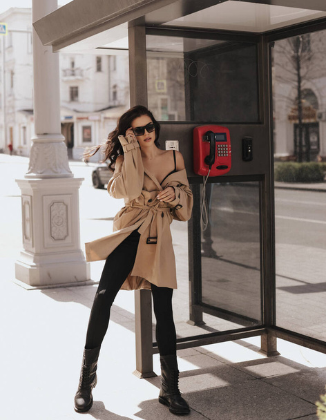 fashion outdoor photo of beautiful woman with dark hair in elegant beige coat and boots posing in telephone booth on a city street - Photo, image