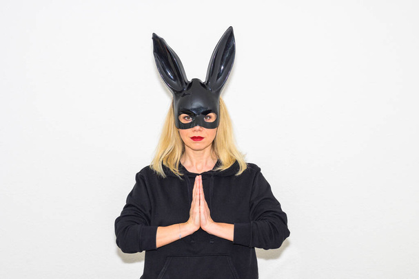 Sensuous blond woman wearing a black bunny mask standing with steepled hands looking at the camera with a quiet smile over white - Valokuva, kuva