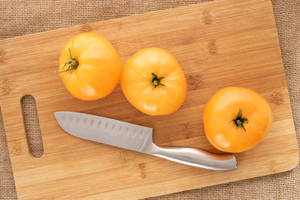 Three organic yellow tomatoes with a metal knife on a wooden cutting board on jute fabric, close-up, top view. - Photo, Image