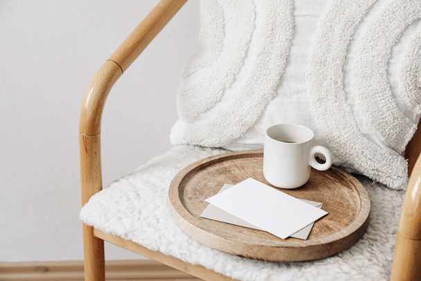 Trendy Scandinavian interior still life. Breakfast concept. Blank greeting card, invitation and cup of coffee on wooden tray. Stationery mock-up. Wooden chair, wool teddy armchair, white cushion. - Photo, Image