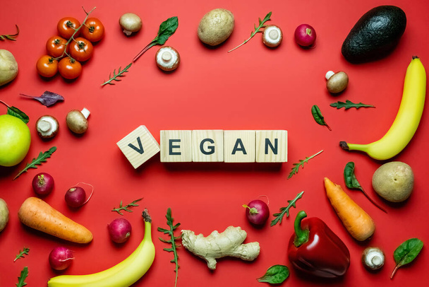 Bandeja leigos com letras vegan em cubos de madeira perto de vegetais maduros e frutas no fundo vermelho  - Foto, Imagem