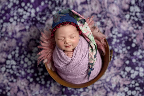 Pequena menina ucraniana recém-nascido dorme pacificamente em uma cesta em um fundo roxo. Em sua cabeça um chapéu de flores. - Foto, Imagem
