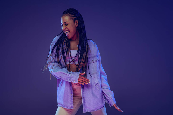 Emotional african girl in jeans and shirt dancing, having fun isolated on dark blue background. Concept of beauty, art, fashion, youth and emotions - 写真・画像