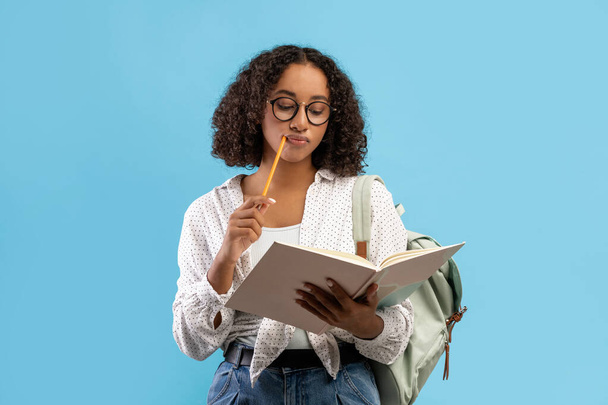 Προνοητική Αφροαμερικανίδα φοιτήτρια διαβάζει βιβλίο, σκέφτεται πάνω από το σπίτι ανάθεση σε μπλε φόντο στούντιο - Φωτογραφία, εικόνα