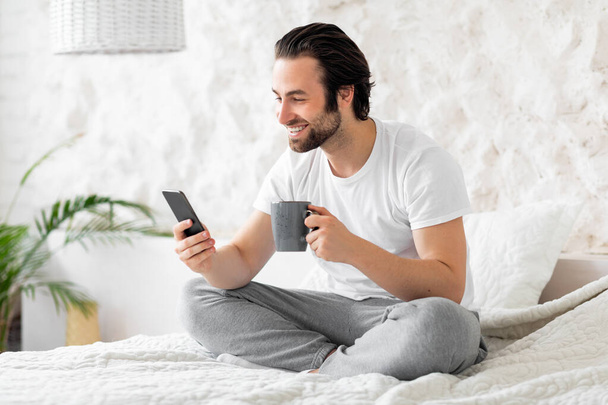 Ξεκούραστος όμορφος άντρας που αράζει στο κρεβάτι στο σπίτι, χρησιμοποιώντας smartphone - Φωτογραφία, εικόνα