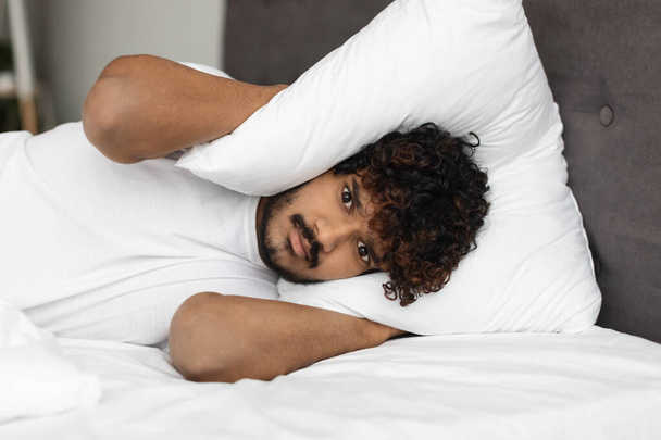 Irritated eastern man laying in bed, covering head with pillow - Foto, afbeelding