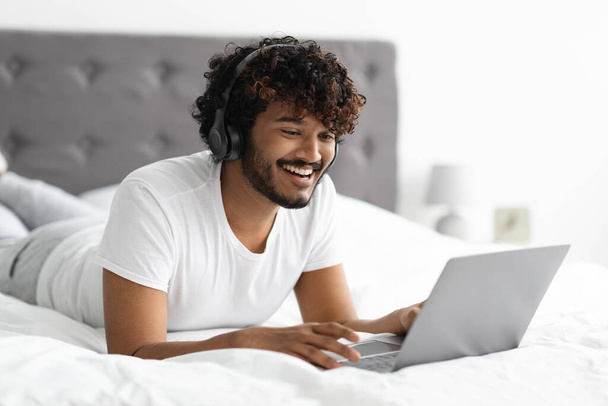 Relaxed eastern guy enjoying weekend in bed, using laptop - Photo, Image