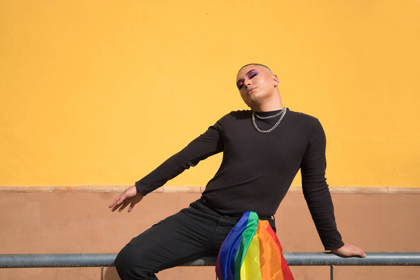 Persona no binaria, joven sudamericano, fuertemente maquillada, sentada en una barandilla con una bandera de orgullo gay sostenida por un bolsillo de pantalón. Concepto reina, lgbtq +, orgullo, queer. - Foto, imagen