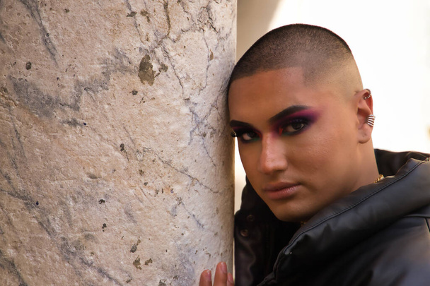 Portrait of non-binary person, young and South American, heavily make up and with serious gesture leaning on a marble column. Concept queen, lgbtq+, pride, queer. - Foto, Imagem