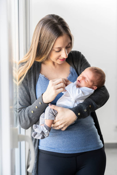 Νεαρή μητέρα με το νεογέννητο μωρό της στην αγκαλιά της στο σπίτι κοντά στο παράθυρο - Φωτογραφία, εικόνα