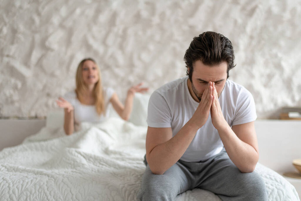 Young man feeling depressed, sitting on bed at home, his angry wife shouting at him, free space. Family problems concept - Foto, Imagen