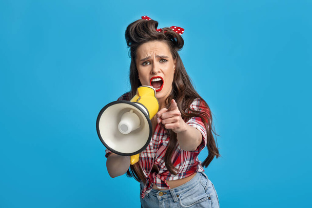 Agitated pinup woman in retro style wear shouting into megaphone, pointing at you on blue background - Fotoğraf, Görsel