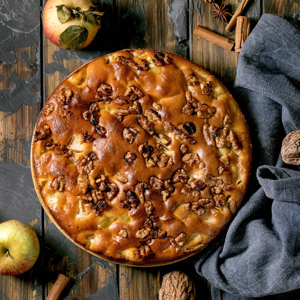 Homemade apple and walnuts cake Charlotte on plate with fresh gardening apples, cinnamons and nuts around over dark wooden plank background. Autumnal home baking. Flat lay. Square image - Φωτογραφία, εικόνα