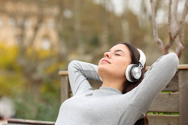 Adolescente rilassato che si riposa ascoltando musica con le cuffie in una panchina in un parco - Foto, immagini