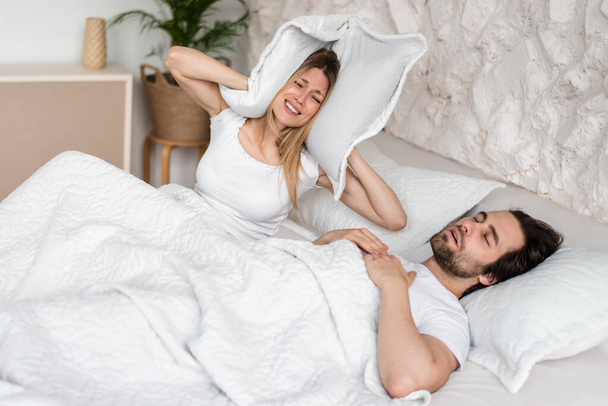 Ronquidos chico joven y su novia frustrada cubriendo las orejas con almohada en la cama en casa - Foto, imagen
