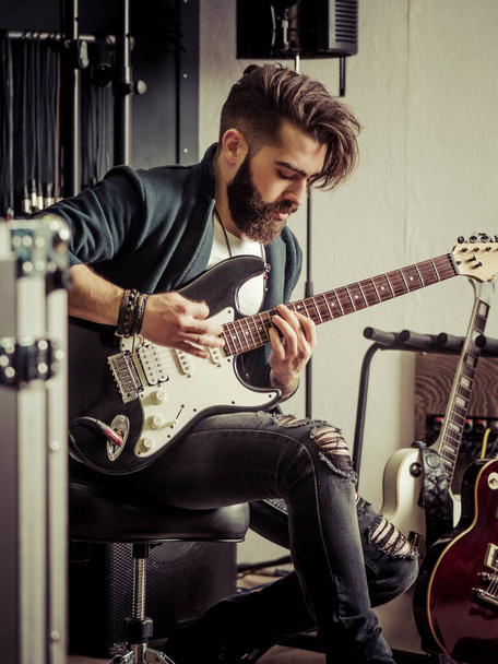 Young man with beard sitting and playing his electric guitar in a recording studio. - Photo, image