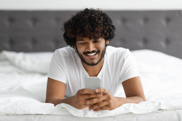 Portrait of handsome hindu guy laying on bed with phone - Foto, Imagem