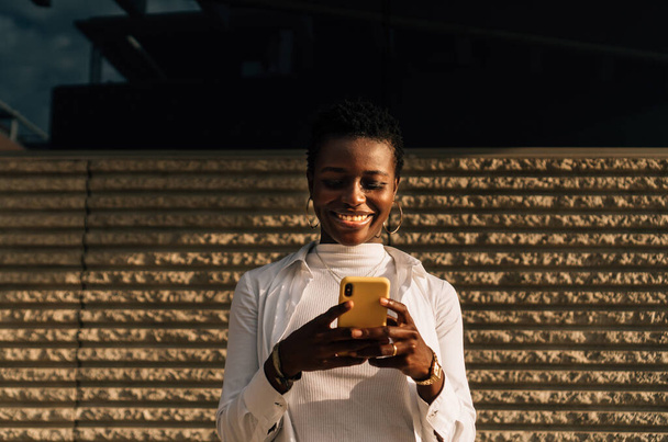 Αφροαμερικανή γυναίκα χαμογελά ενώ χρησιμοποιεί το κινητό της τηλέφωνο σε εξωτερικούς χώρους φωτισμένο με φυσικό φως. - Φωτογραφία, εικόνα