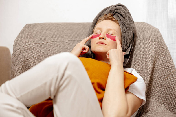Young woman with casual clothes, towel on head and eye patches sitting on couch and resting. Relax after taking shower. Cosmetic for women, skin care. Self care, time for yourself. Relaxing at home. - Photo, Image