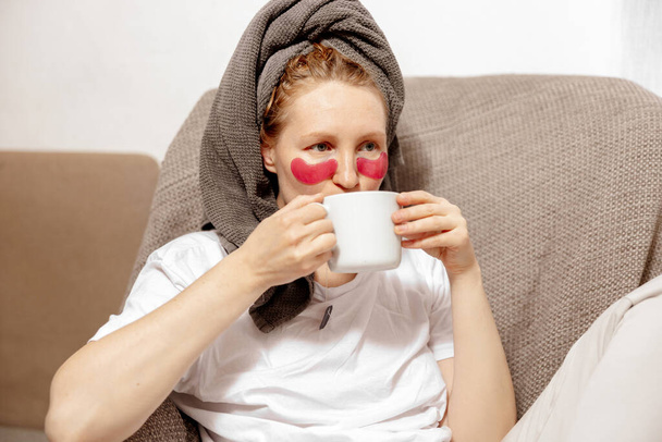 Young woman with casual clothes, towel on head and eye patches drinking hot tea and resting. Relax after taking shower. Cosmetic for women, skin care. Self care, time for yourself. Relaxing at home. - Foto, Imagen