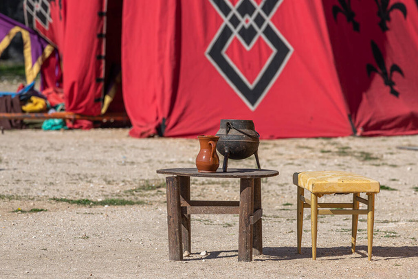 closeup of a wooden table and chair with an old cauldron and clay jug on the table as a decoration in the recreation of a military camp from medieval times - Photo, Image