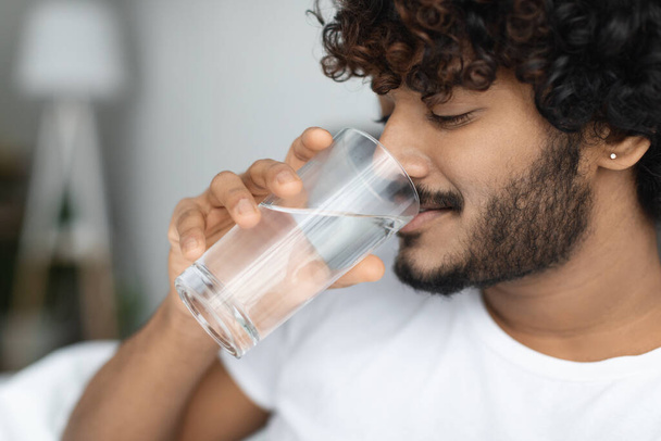 Happy curly young man taking glass of water in bed - Photo, image