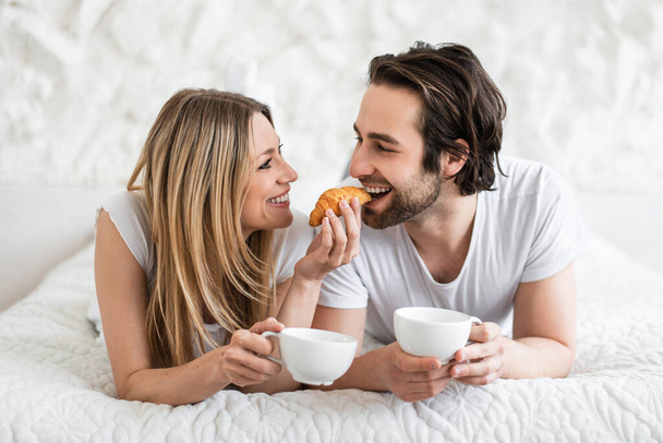 Loving young couple eating breakfast and drinking coffee in bed at home. Lazy romantic morning concept - Фото, изображение