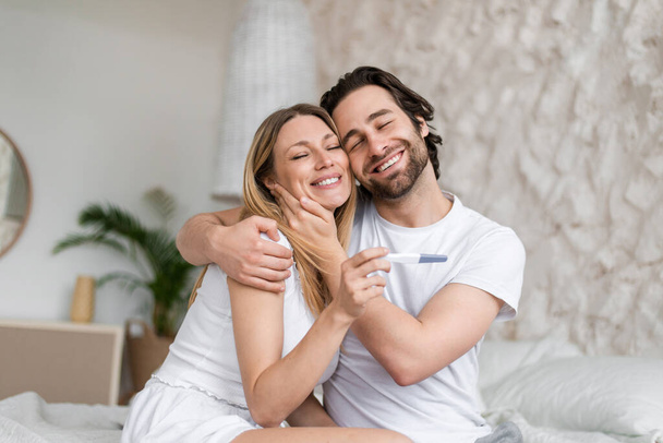 Feliz pareja joven celebrando el resultado positivo de la prueba de embarazo, abrazos y abrazos en la cama en casa - Foto, Imagen