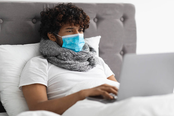Sick young man in face mask staying in bed, using laptop - Fotó, kép