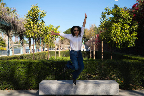 young and beautiful south american girl jumps from a park bench while having fun and smiling. Concept physical activity, fun, happiness, celebration. - Foto, Imagen