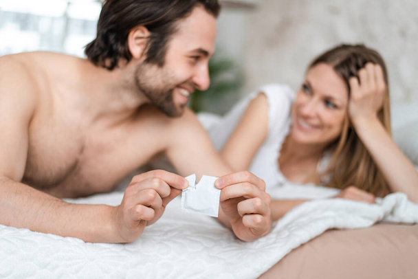 Happy young couple lying in bed and opening condom, selective focus. Pregnancy and STD control concept - Foto, Imagem