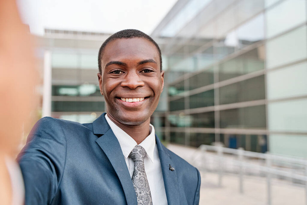 młody afrykański biznesmen ubrany w formalny garnitur robi selfie poza centrum pracy uśmiechnięty - Zdjęcie, obraz