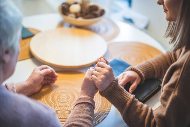 Elder woman with her daughter holding hands together and praying at the table - Фото, изображение