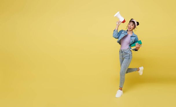 Щаслива азіатська дівчинка, що тримає книжку і стрибає на оголошений мегафон на жовтому тлі. знову до шкільної концепції - Фото, зображення