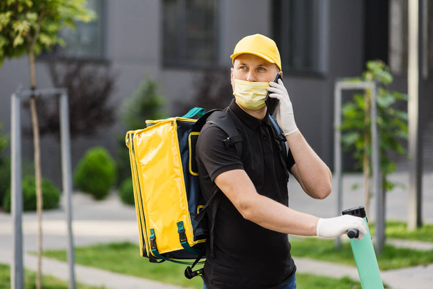 Delivery man in yellow uniform stands with smartphone against background of an apartment building - 写真・画像