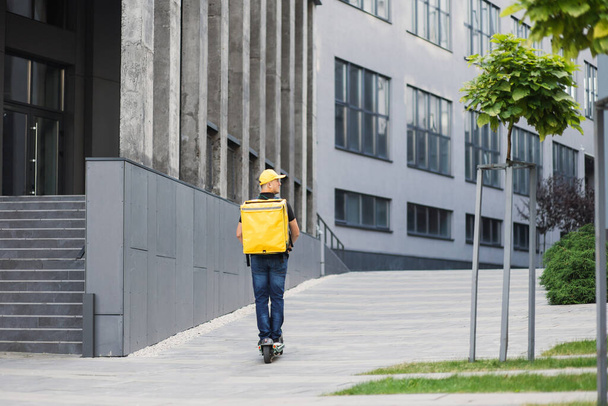 Young man with yellow backpack on his back in casual summer clothes is movingon an electric scooter - Foto, immagini