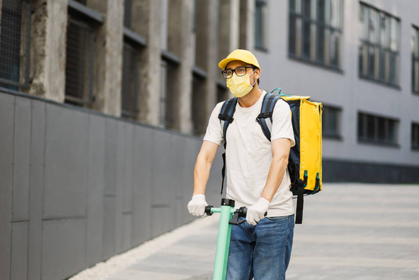 Man courier in yellow face mask on scooter in town streets with a hot food delivery - 写真・画像