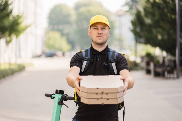 Smiling happy courier holding coffee, pizza box, wearing black t-shirt on the city background. - Fotoğraf, Görsel