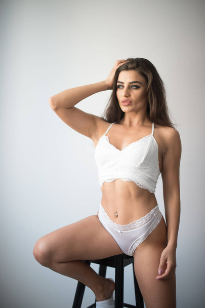 Attractive sexy brunette in white lingerie  posing provocatively, in front of a wall ,studio shot. Portrait of sensual woman with long hair, white bikini and bra in classic boudoir scene in studio - Foto, afbeelding