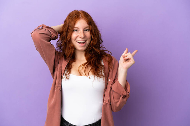 Teenager rothaarige Mädchen über isolierten lila Hintergrund überrascht und zeigt mit dem Finger zur Seite - Foto, Bild