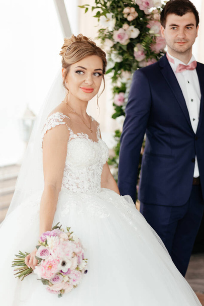 A young beautiful bride in a white dress and the groom stand near the flower arch and hold a large wedding bouquet. High quality photo - Fotografie, Obrázek