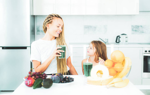 Mother and daughter drinking green smoothie in a white kitchen. Family mother and child daughter blended green and orange smoothie with healthy fruits ingredients. Detox smoothie and healthy food - Foto, Imagem