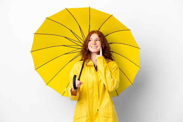 Nastoletnia ruda dziewczyna płaszcz przeciwdeszczowy i parasol izolowany na białym tle myśląc pomysł podczas patrząc w górę - Zdjęcie, obraz