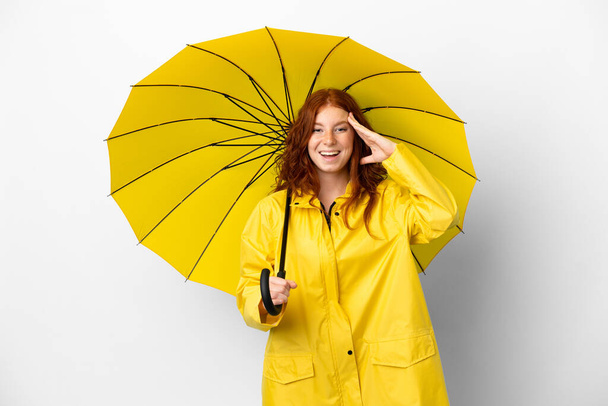 Teenager redhead girl rainproof coat and umbrella isolated on white background with surprise expression - Fotó, kép